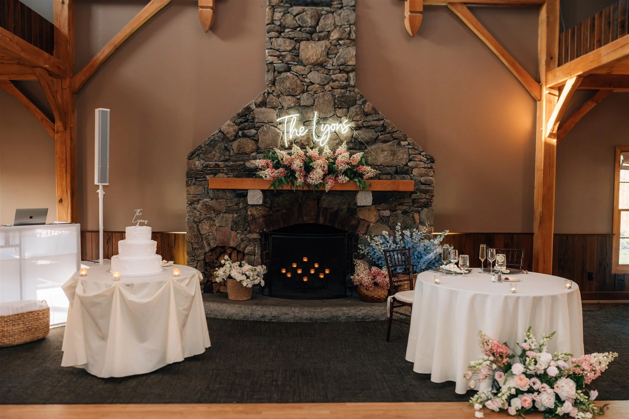 Wedding reception setup in a rustic room with a stone fireplace, floral arrangements, a neon sign reading 'The Lyons' above the fireplace, a three-tiered wedding cake, and tables set for dining with candles and floral decor.