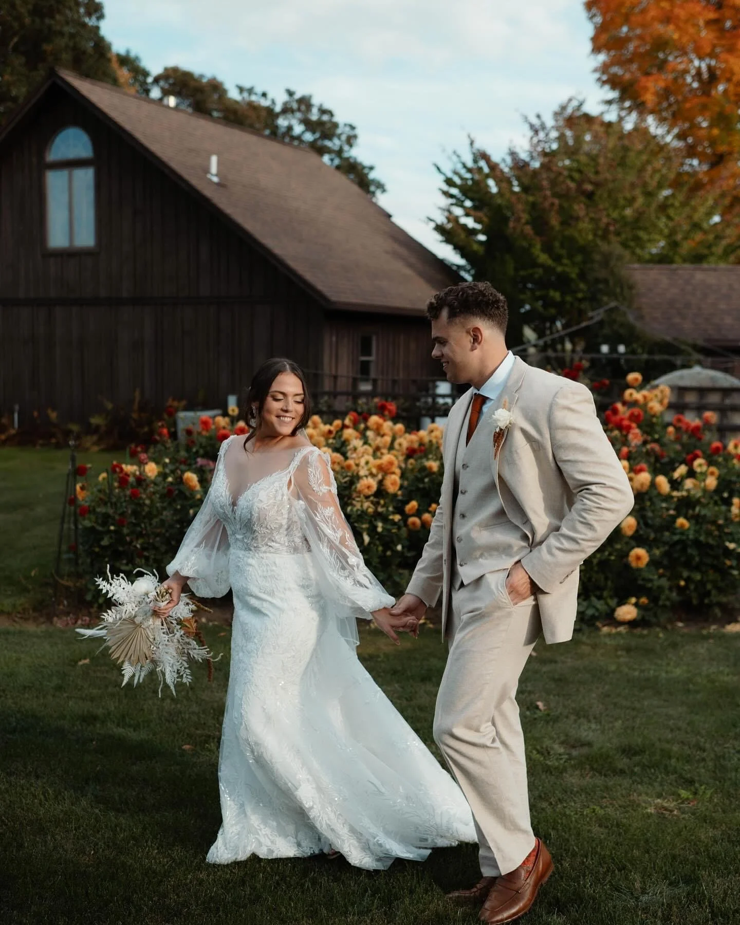 A bride and groom holding hands and smiling in a garden with blooming flowers and a wooden barn in the background.