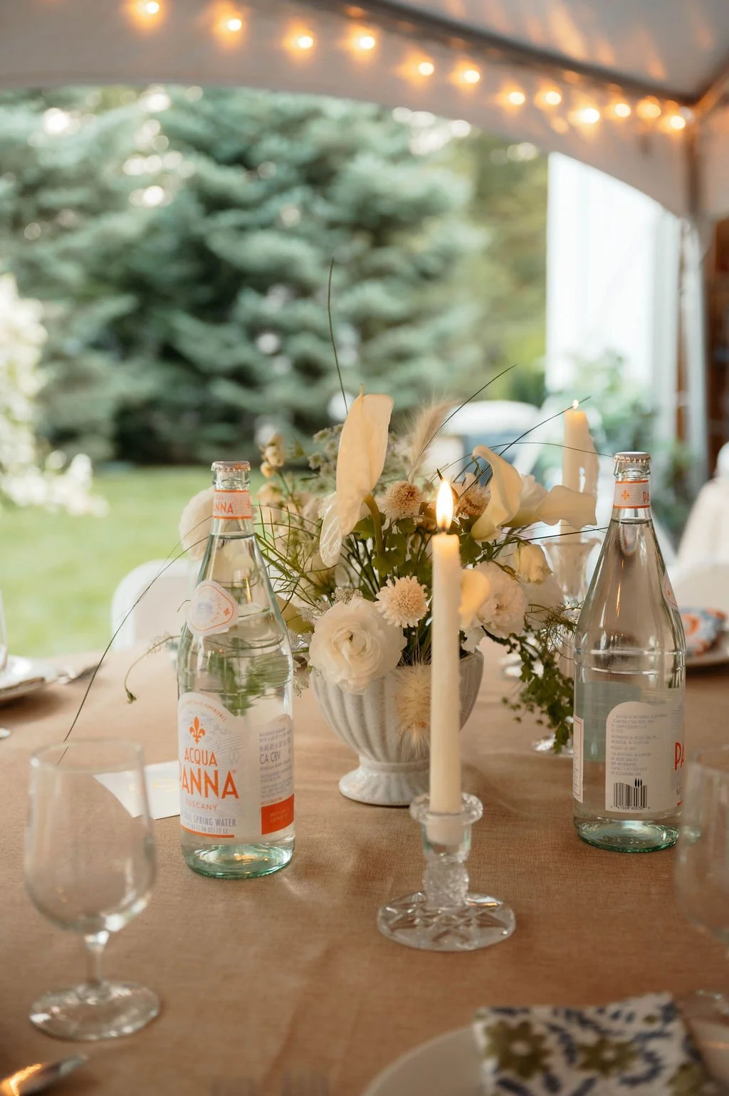 A table decorated with a bouquet of white flowers, a lit candle, and bottles of sparkling water, set under a canopy with string lights, overlooking a green outdoor area.