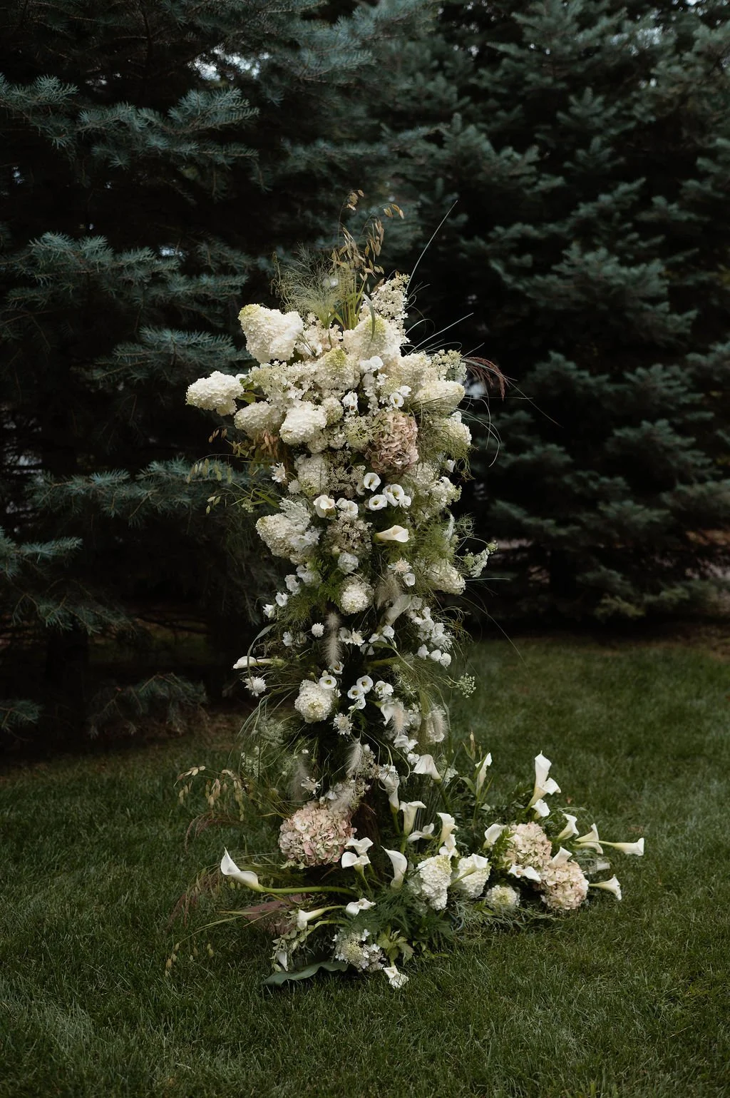 A large white floral arrangement with hydrangeas, calla lilies, and other white flowers, set outdoors on grass with pine trees in the background.