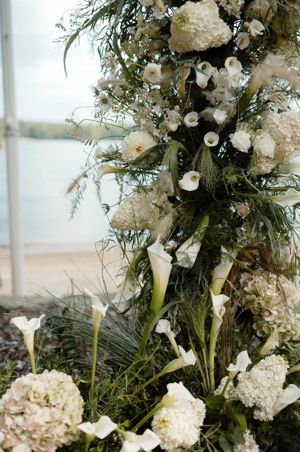 A large floral arrangement with white flowers, including calla lilies, hydrangeas, and roses, against a backdrop of a lake and a partly cloudy sky.