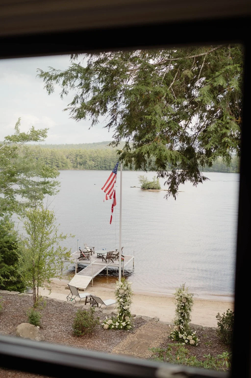 A view of a lakeside scene through a window frame, showing a small dock with Adirondack chairs, an American flag, and floral arrangements at the shoreline, surrounded by trees.