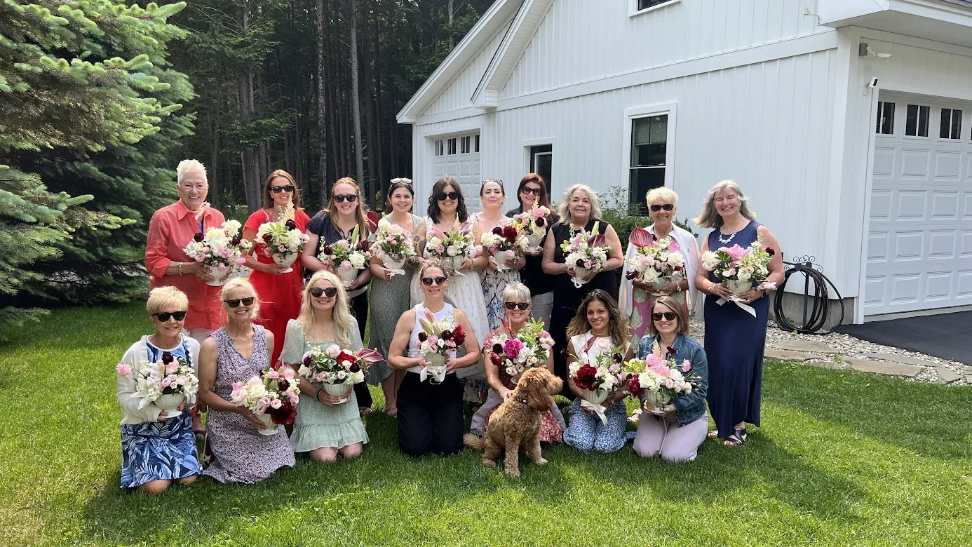 Group of women posing outdoors in front of a white house, holding bouquets of flowers, with a dog and a background of trees.