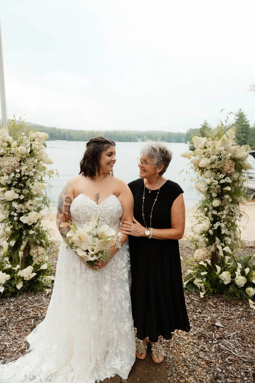 A bride and an older woman, possibly her mother, smiling at each other during a wedding ceremony outdoors near a lake with floral arrangements on either side.