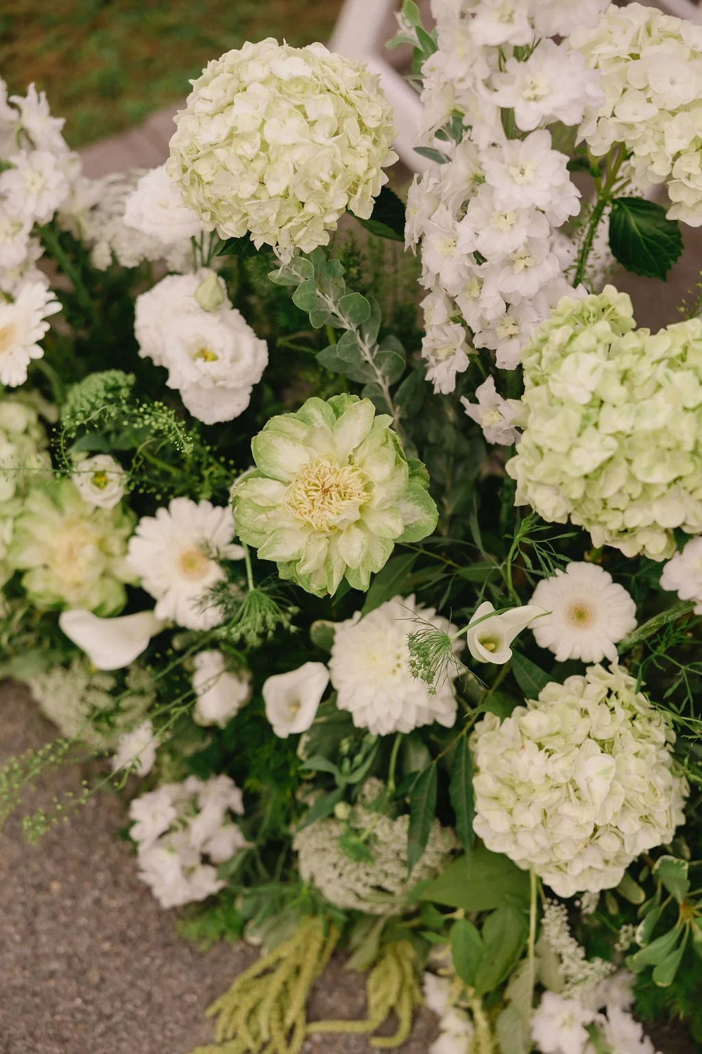 A bouquet of white and cream flowers, including hydrangeas, roses, and daisies, arranged on a gravel surface.
