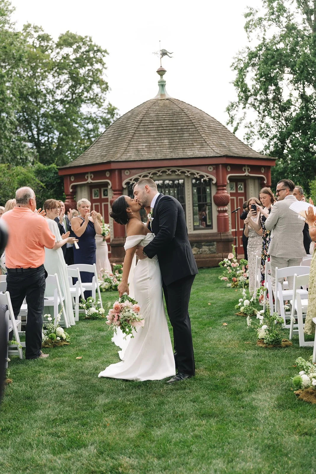 A bride and groom sharing a kiss at their outdoor wedding ceremony, surrounded by seated guests and floral decorations, with a small ornate building in the background.