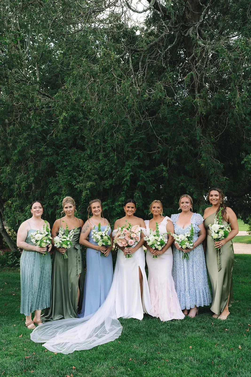 A group of seven women dressed in colorful bridesmaid dresses standing outdoors on grass, holding bouquets of flowers, with a large tree in the background.