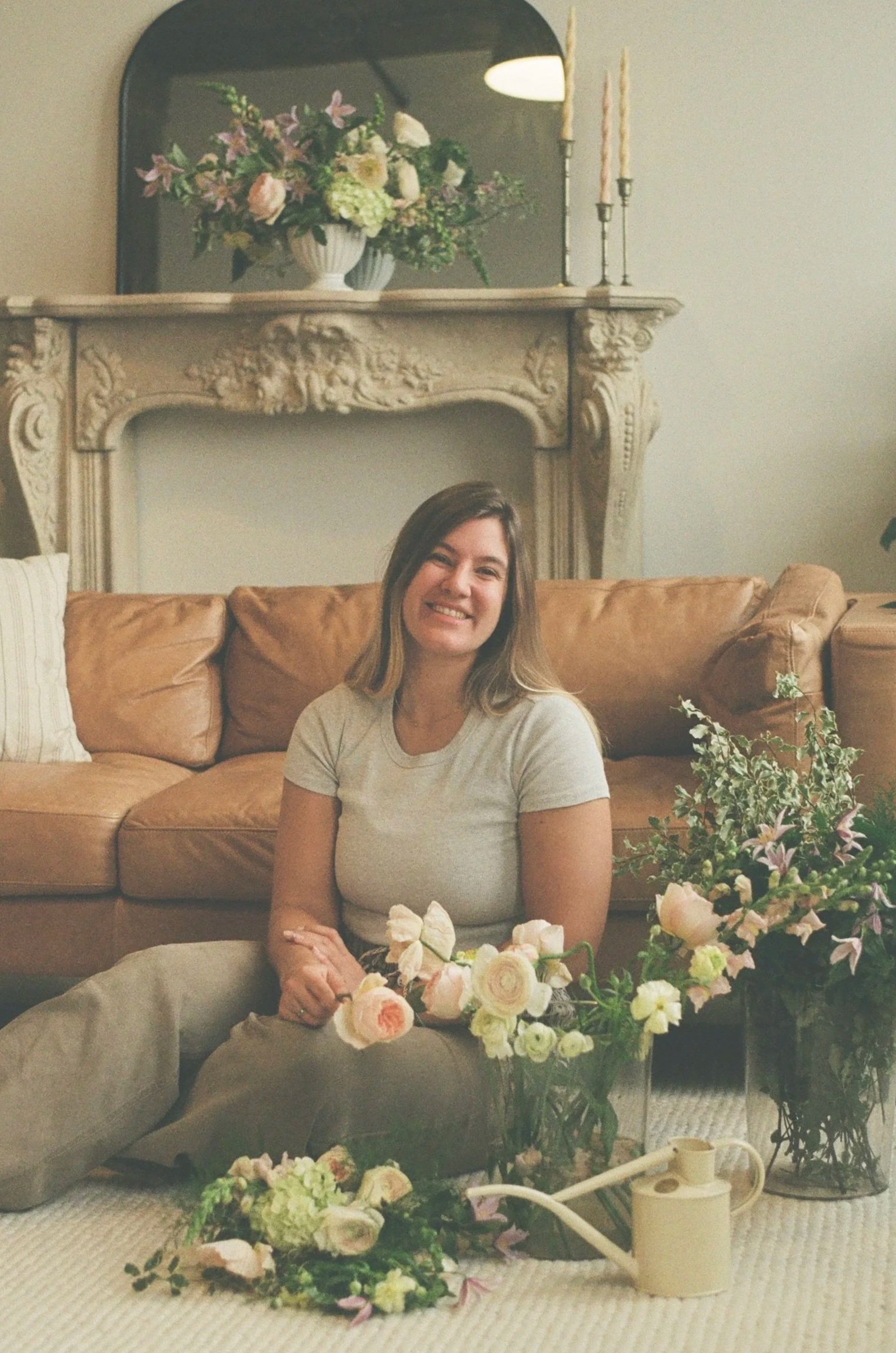 A woman sitting on the floor in front of a brown leather couch, surrounded by various flowers and a watering can, smiling at the camera in a living room with a decorative fireplace and a mirror above it.