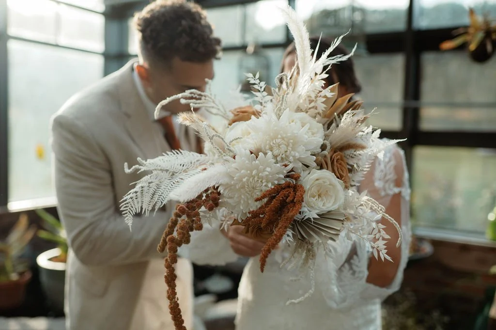 A wedding couple is holding a large bouquet with white and beige flowers, dried grasses, and decorative foliage, inside a glass greenhouse.