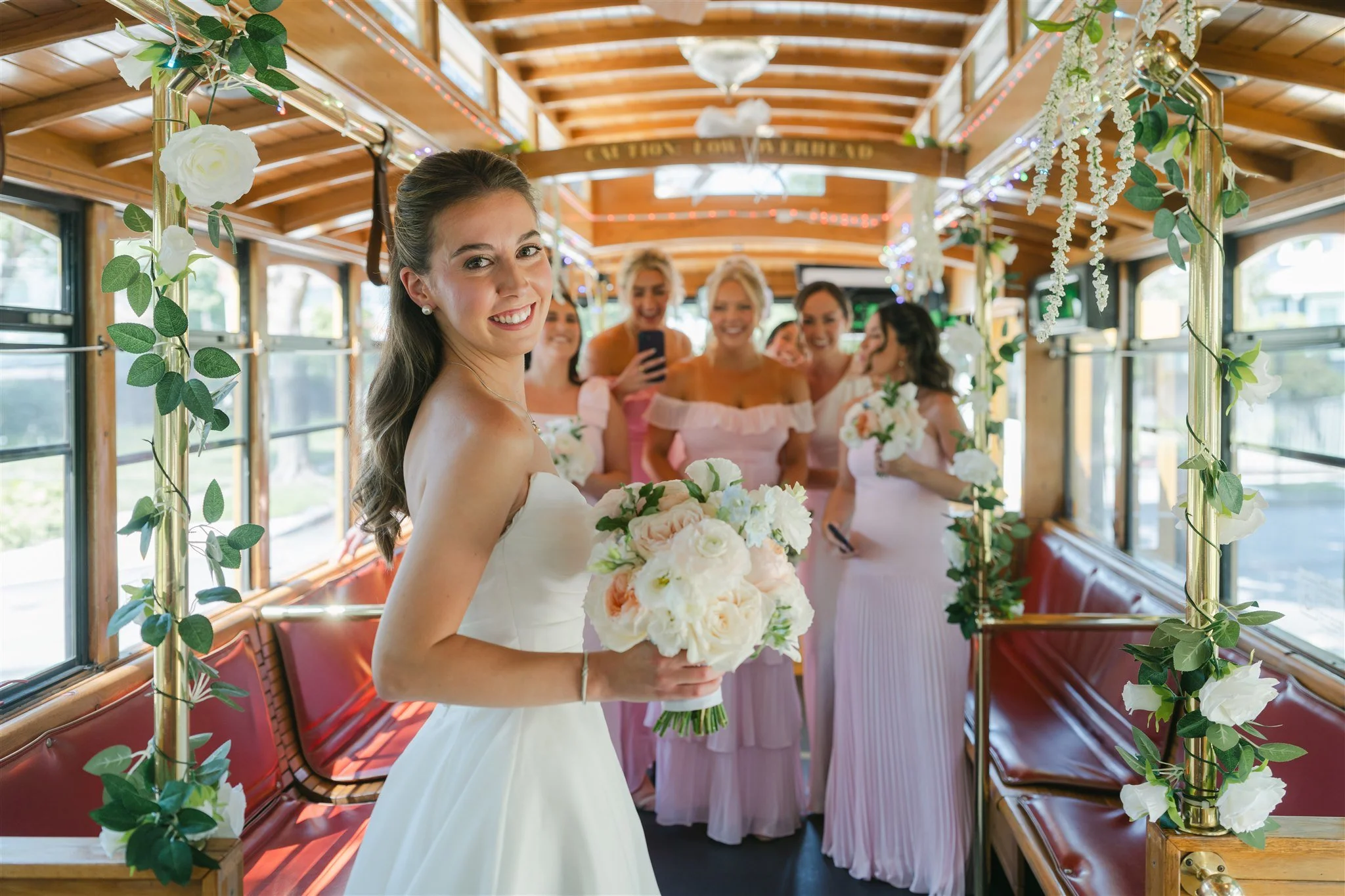 A bride smiling and holding a bouquet of white and peach flowers inside a decorated trolley, with her bridesmaids behind her taking a selfie, all dressed in pastel colors.