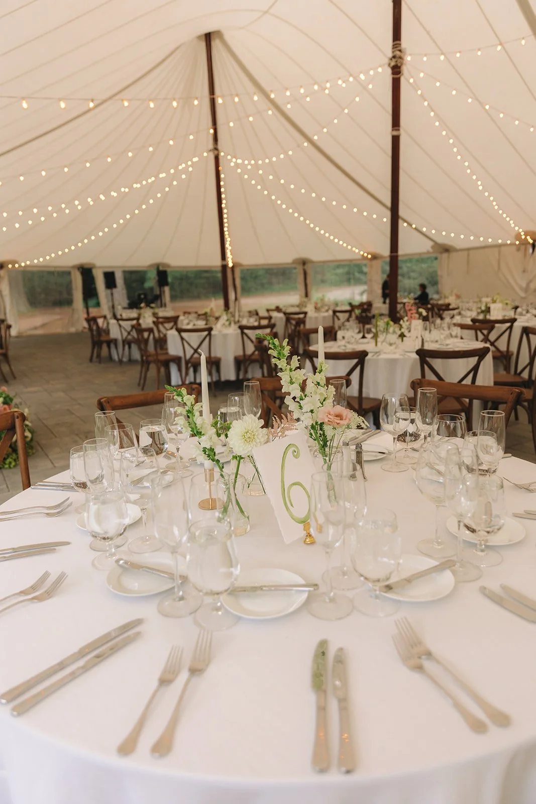 Elegant wedding reception setup inside a large tent with string lights, decorated tables with floral centerpieces, candles, and place settings.