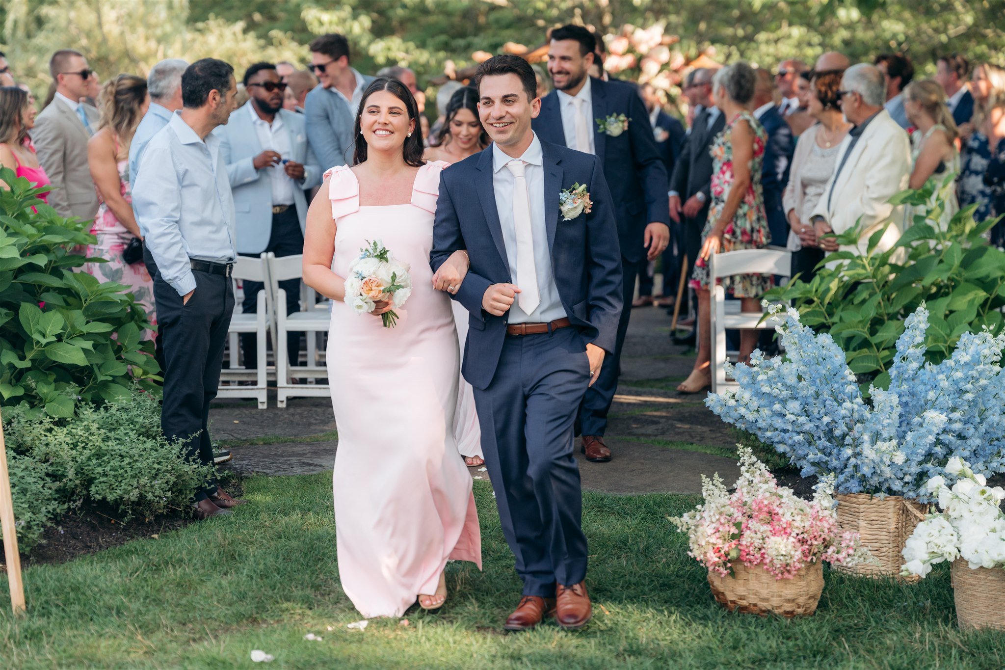 A bride and groom walk arm-in-arm down an outdoor aisle at their wedding, surrounded by seated guests and floral arrangements.