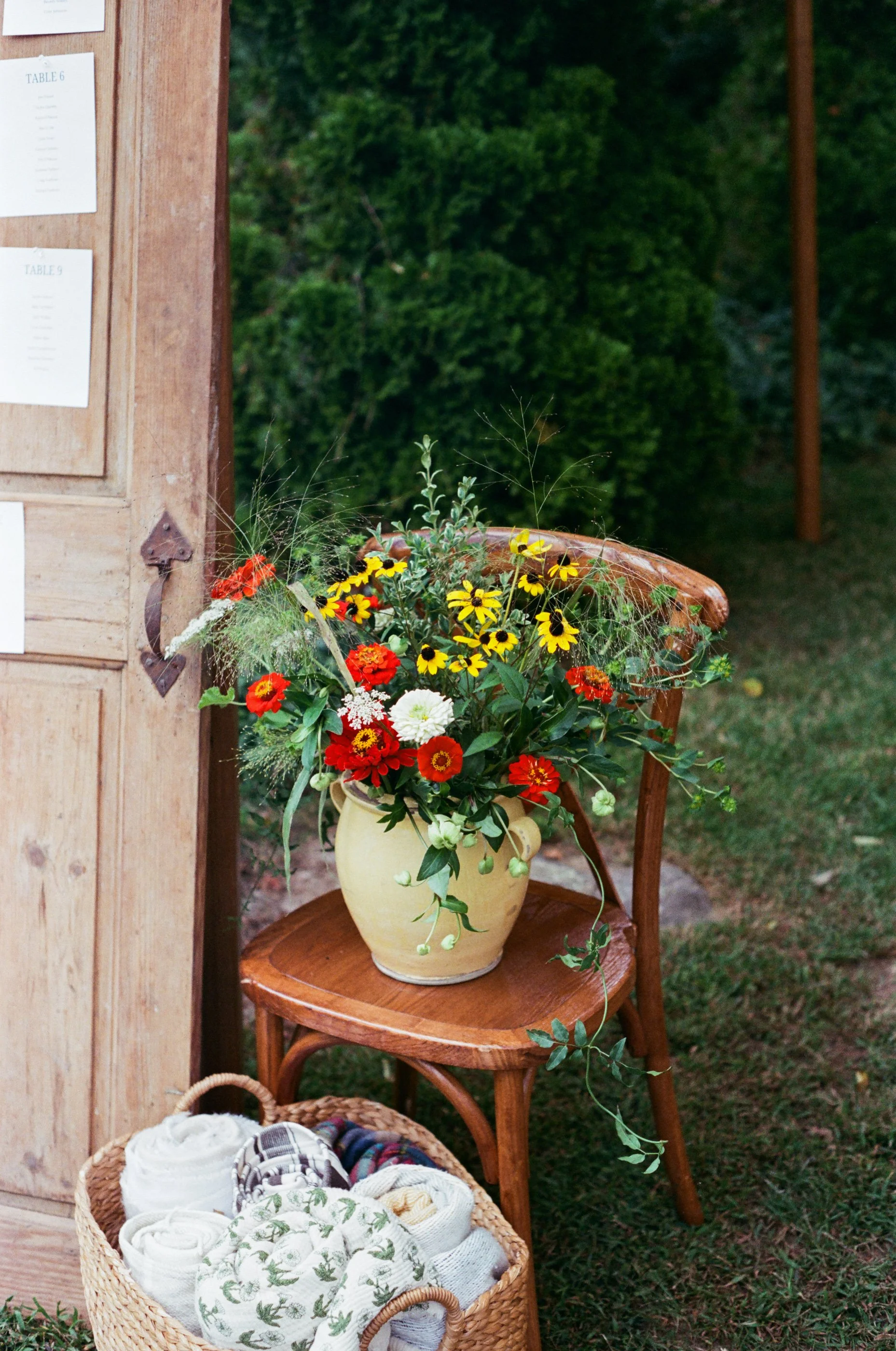 A beige ceramic vase filled with colorful flowers, including yellow, red, white, and green foliage, placed on a wooden chair outdoors. A wicker basket with rolled-up towels and fabric items is in front of the chair, against a backdrop of green bushes