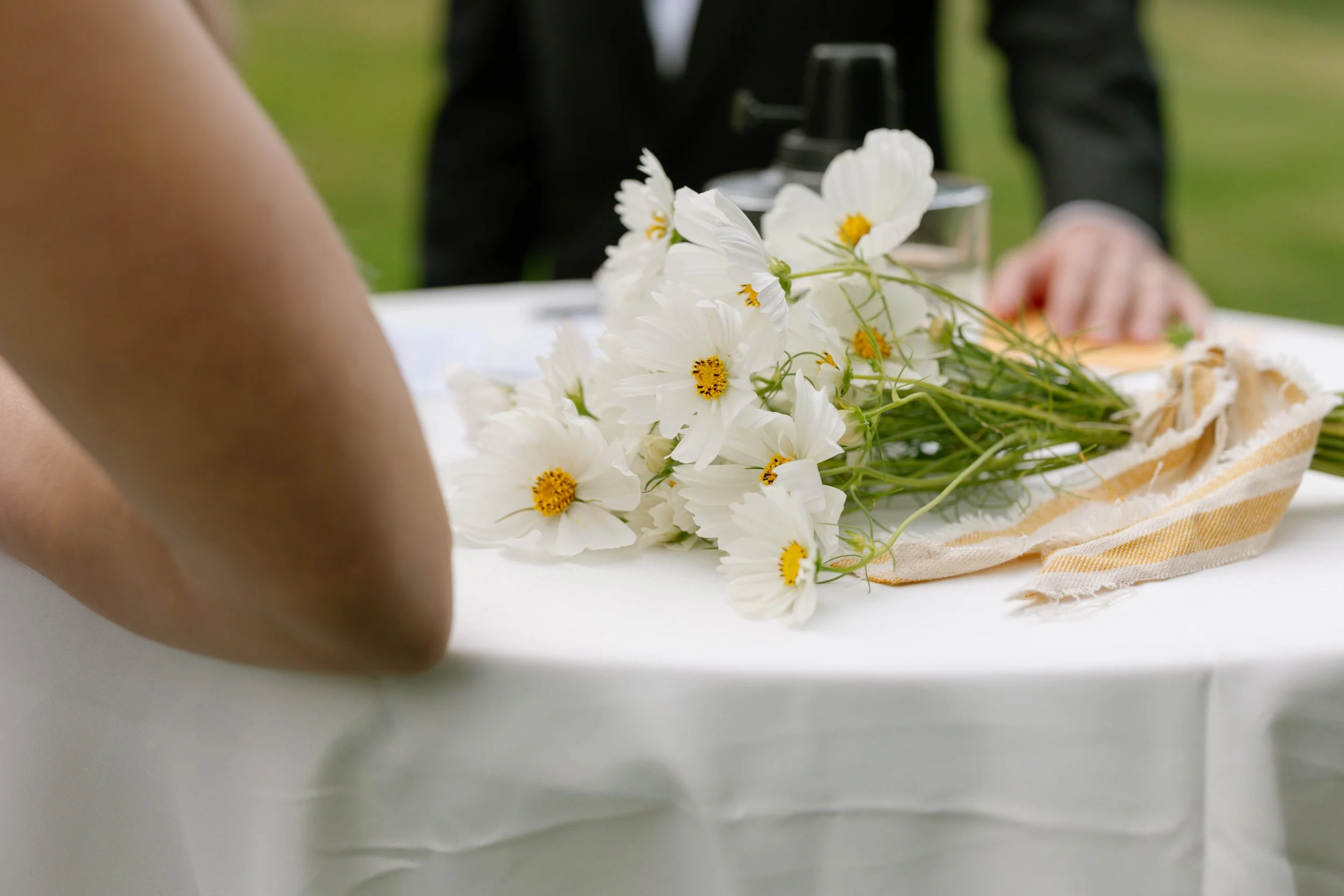 A person sitting at a table with white flowers and a striped cloth. There are some bottles in the background, and a hand is visible in the background.