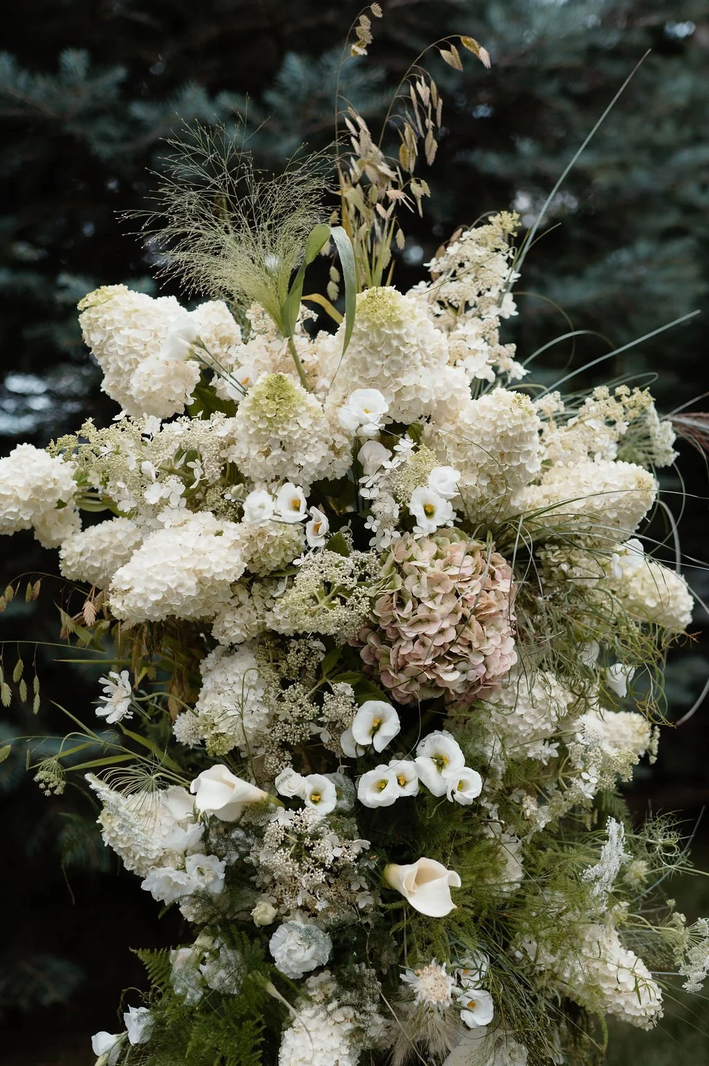A tall bouquet featuring white hydrangeas, calla lilies, and assorted greenery, set against a dark outdoor background.