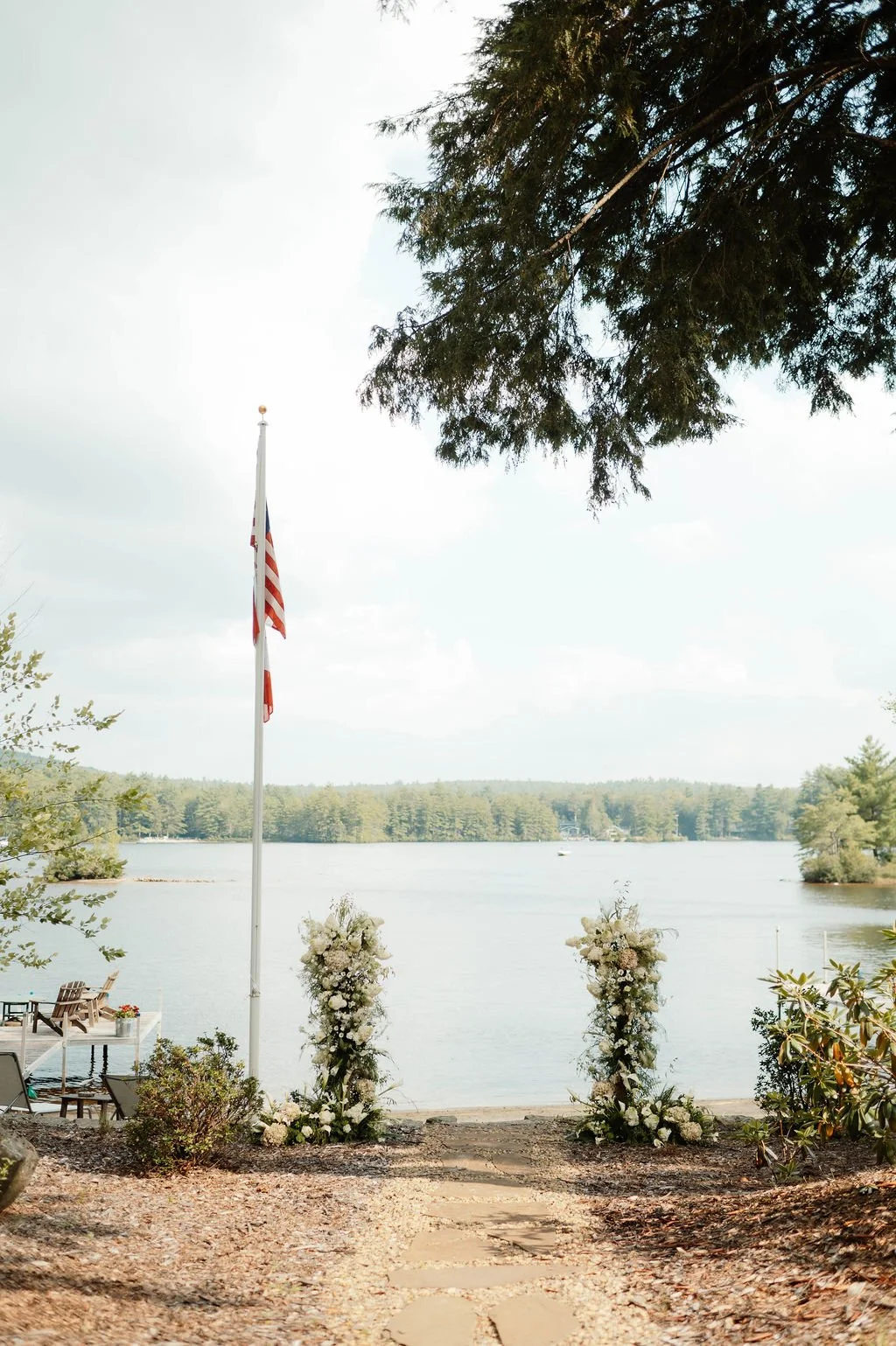 Wedding aisle with floral arrangements leading to a lakeside view, American flag on a flagpole, trees, and outdoor seating.