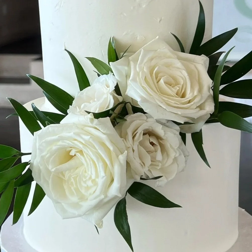 A white floral arrangement with large white roses and green leaves in a white container.