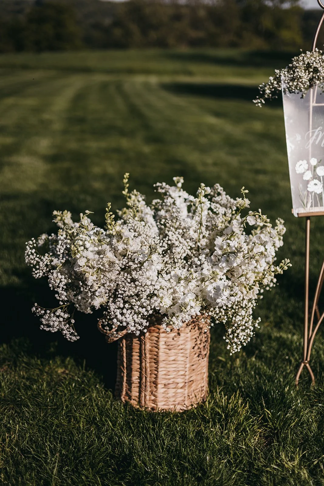 A basket of white flowers on grass, with a blurred outdoor background and a decorative sign on the right.