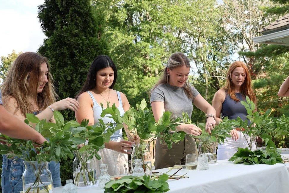 Four women arranging green leafy branches in glass vases on a table outdoors, with trees and a blue sky in the background.