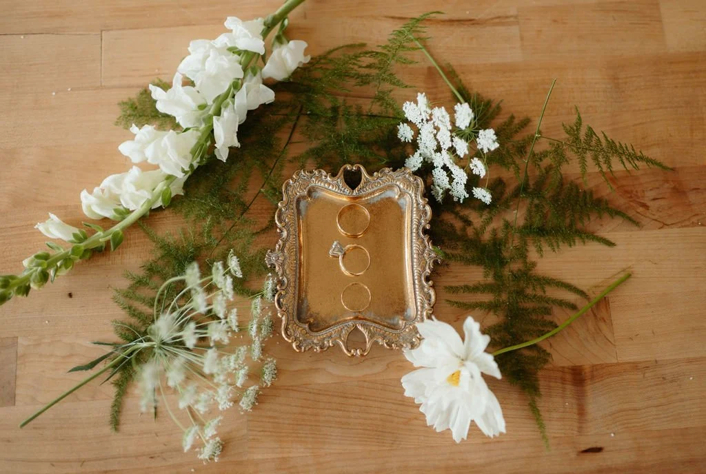 Silver jewelry tray with two rings on it, surrounded by white flowers and green foliage on a wooden surface.