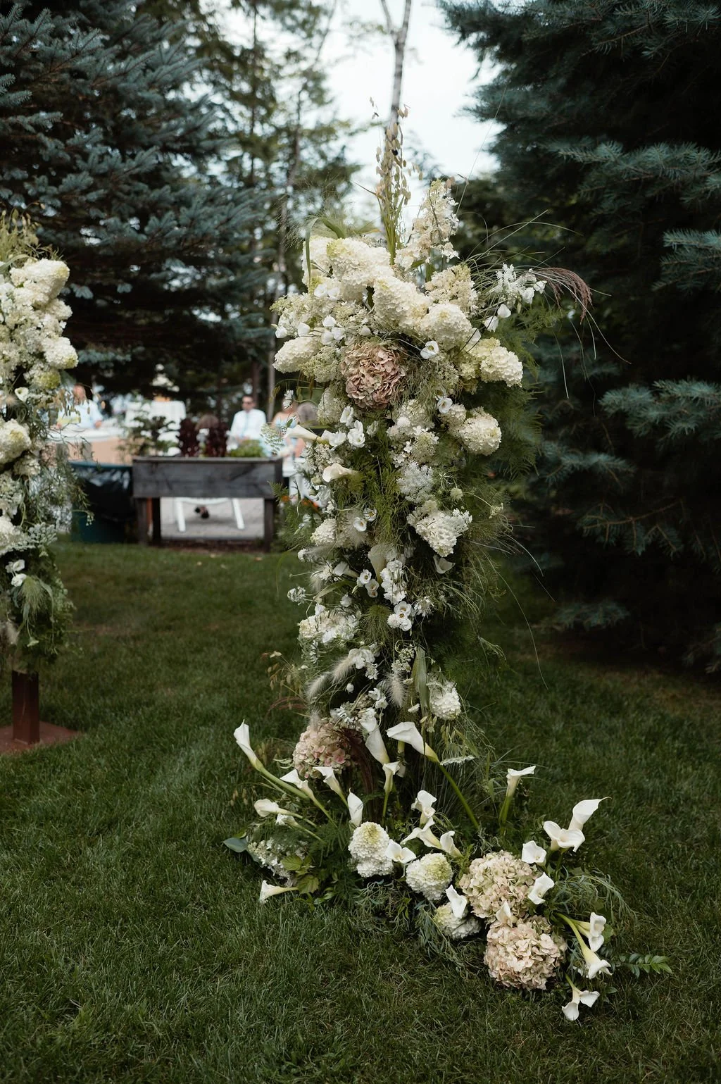 White floral arch with hydrangeas, calla lilies, and other white flowers in an outdoor setting with trees and grass.