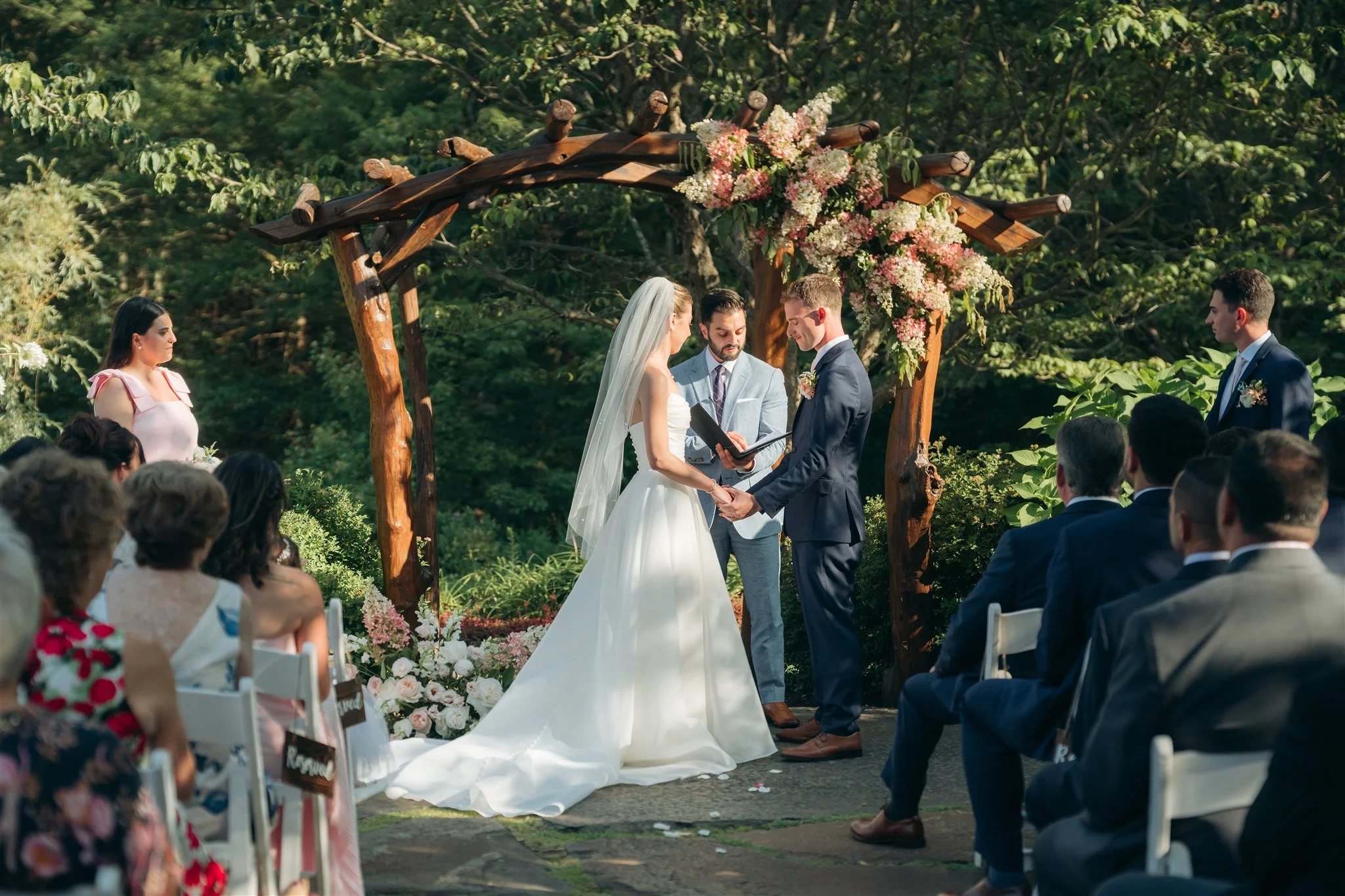 A wedding ceremony outdoors with a bride and groom holding hands, standing under a wooden arch decorated with pink and white flowers, with guests seated on white chairs watching.