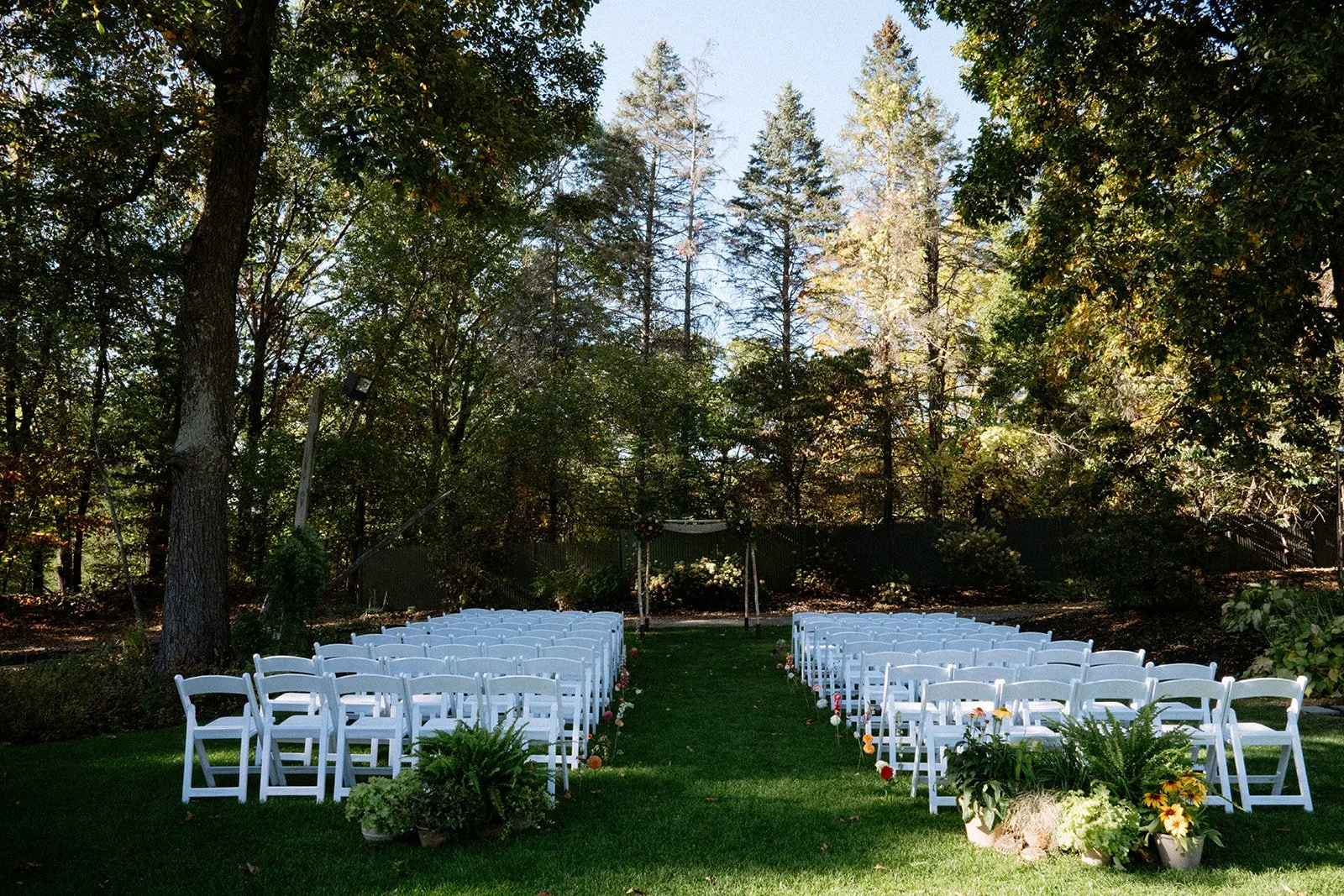 Outdoor wedding ceremony setup with white chairs arranged on either side of a grassy aisle, decorated with flowers, in a wooded area with tall trees.