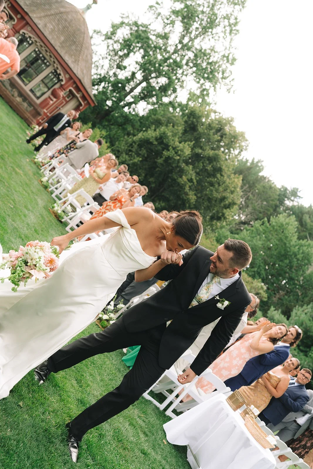 Bride and groom sharing a kiss at an outdoor wedding ceremony with guests watching and greenery in the background.