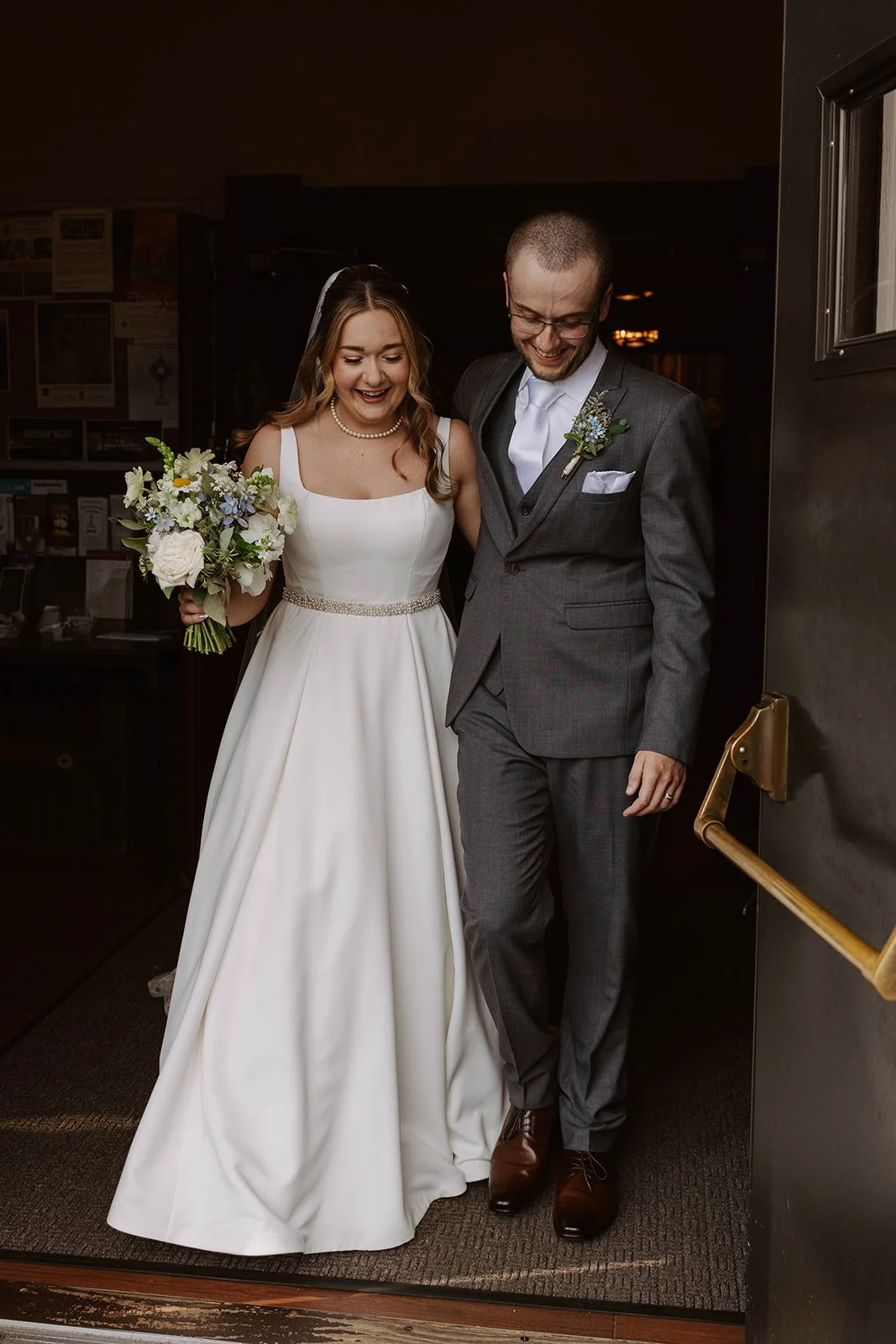 A bride and groom smiling and walking together into a building, with the bride holding a bouquet of flowers and wearing a white wedding dress, and the groom dressed in a gray suit with a boutonniere.