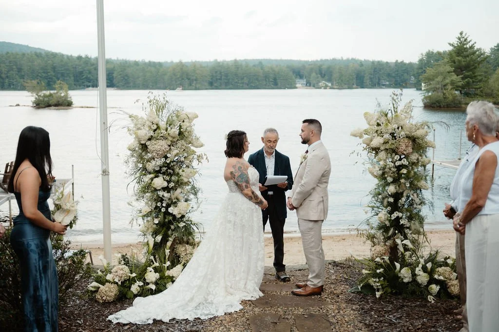 A couple getting married outdoors by a lake, with an officiant and guests around them. The scene is decorated with large floral arrangements, and there are trees and water in the background.