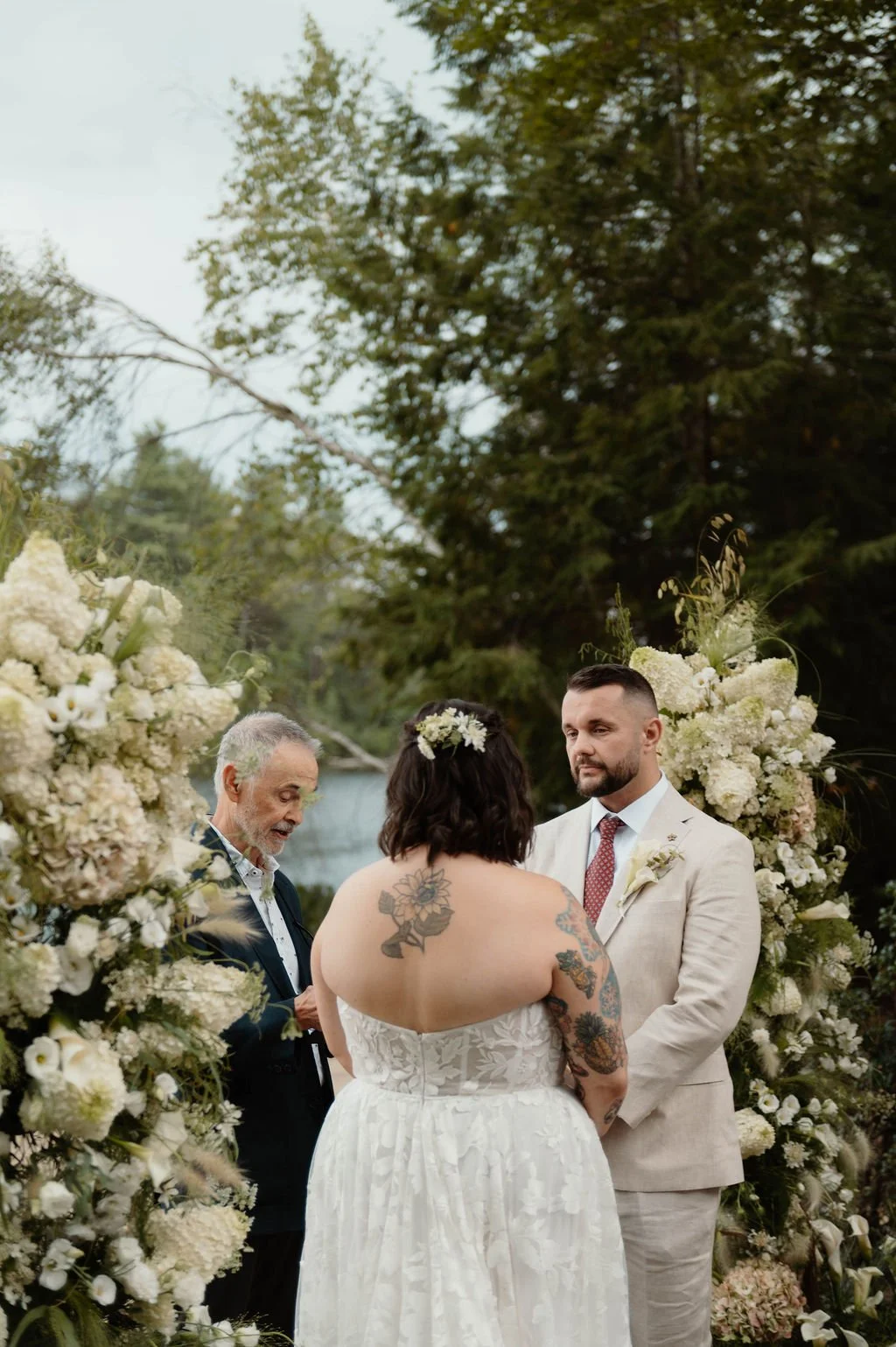 A couple getting married outdoors with a officiant. The bride has tattoos on her back and arms, and the groom is in a light-colored suit. They are standing in front of floral arrangements by a body of water and surrounded by trees.