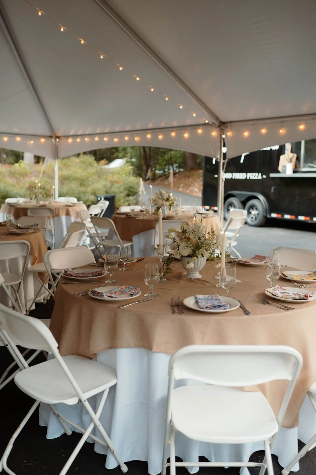 A decorated outdoor event tent with round tables covered with tan tablecloths, set for a meal with place settings, glassware, and floral centerpieces. String lights are hanging inside the tent, and in the background, there is a food truck and greener