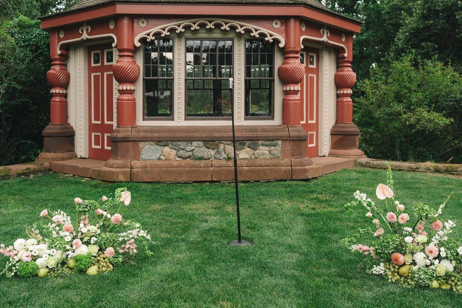 A well-manicured lawn with two floral arrangements on either side. In the background, there is a decorative small building with red and cream details, large windows, and stone accents.