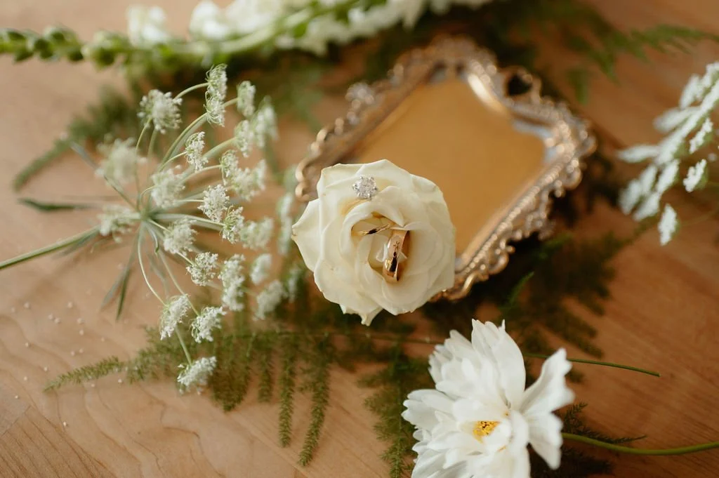 A wooden surface with white flowers, a decorative golden mirror, and two gold rings on a white rose.