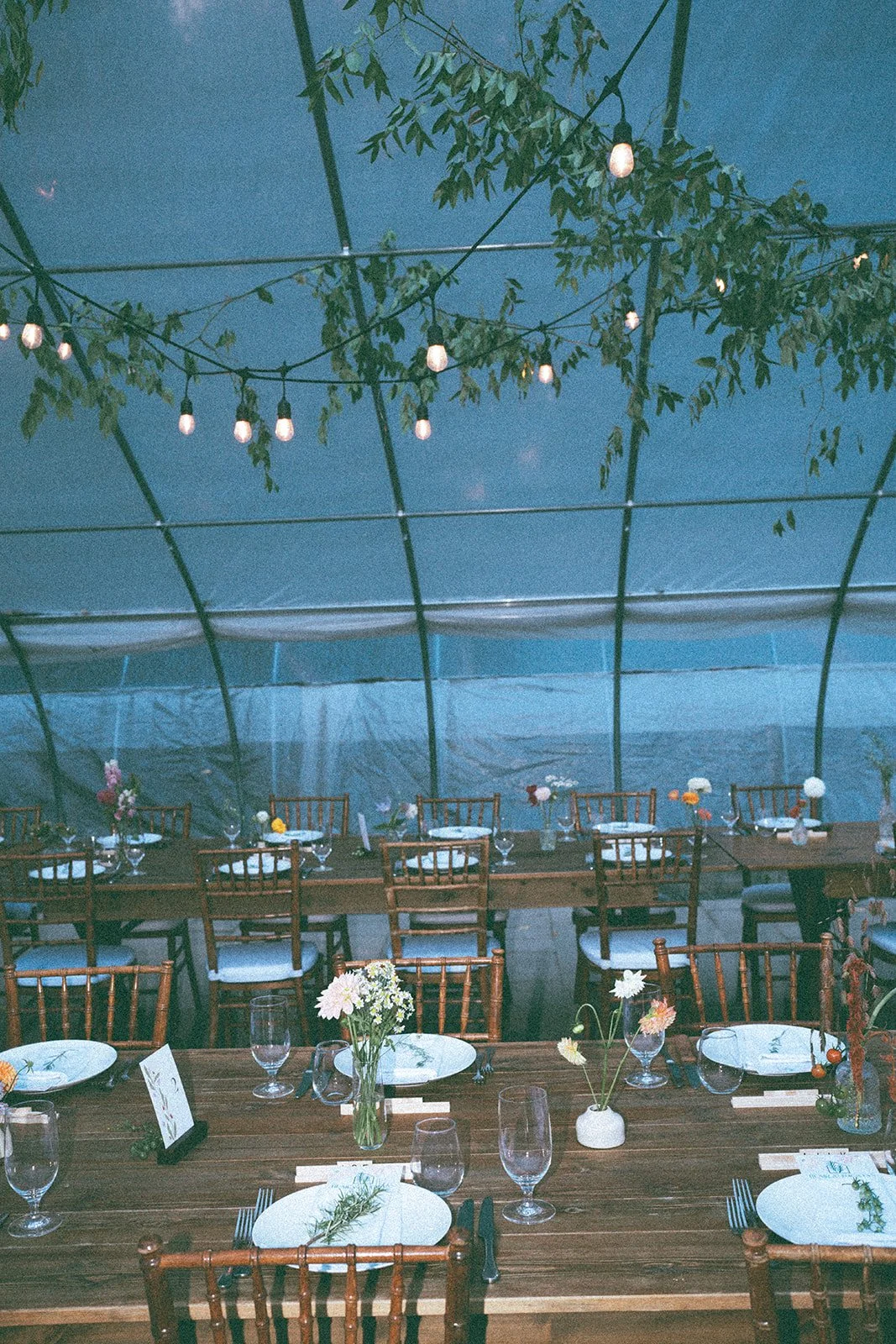 Indoor event space with wooden tables set for a meal, decorated with small flower arrangements and white plates, surrounded by wooden chairs. String lights hang overhead, and a tent-like structure with leaf decorations is visible above.