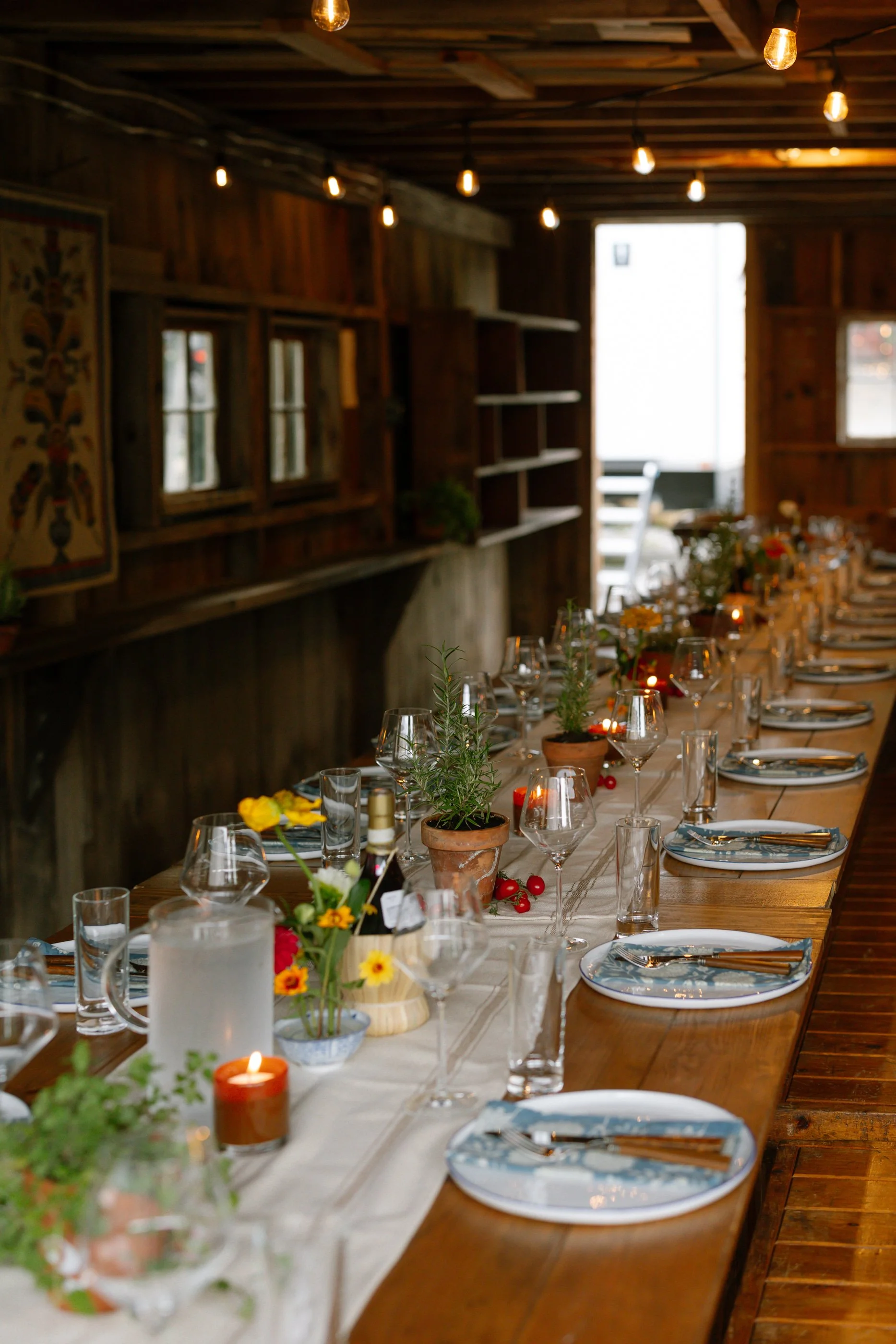 A long wooden dining table set for a meal with plates, silverware, wine glasses, and small potted plants and flowers as centerpieces inside a rustic wooden room with hanging light bulbs.