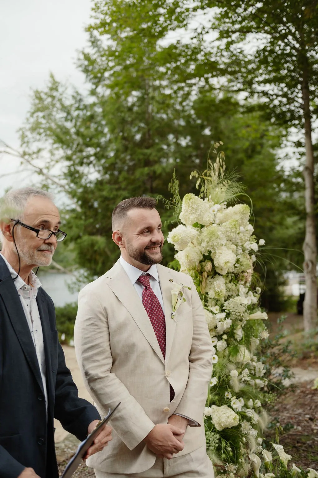 A man in a beige suit with a boutonnière stands outdoors at a wedding ceremony, smiling. Next to him is an officiant in glasses holding a tablet. Behind them is a tall floral arrangement of white flowers and greenery, with a background of trees and a