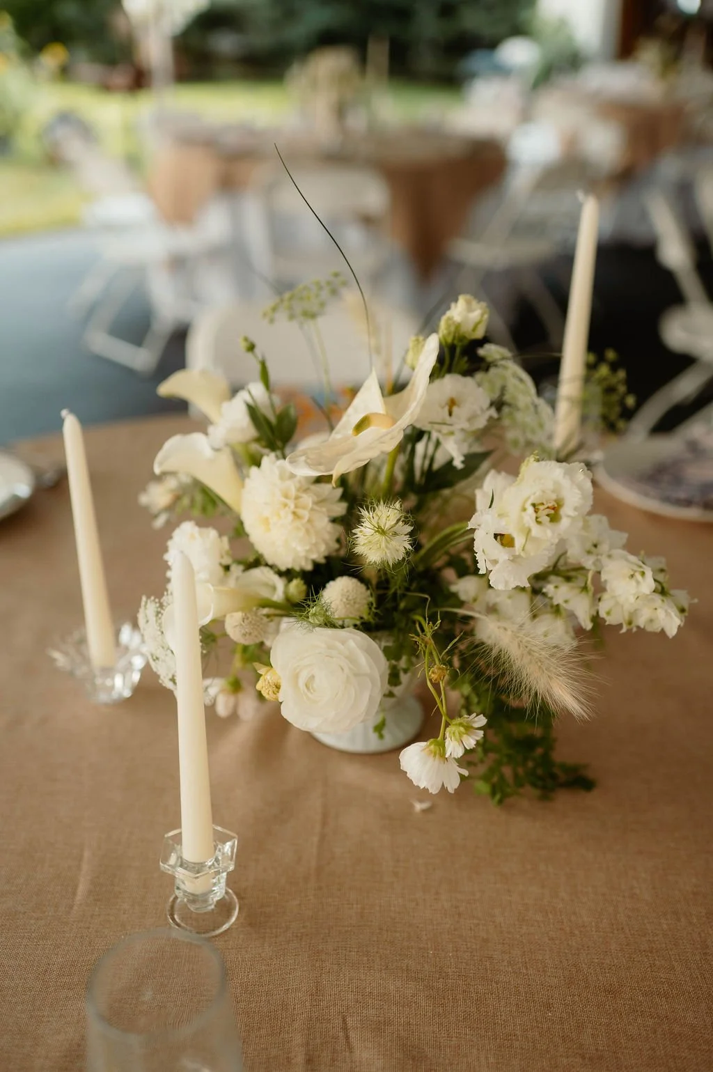 White floral centerpiece with candles on a beige tablecloth at an outdoor event.
