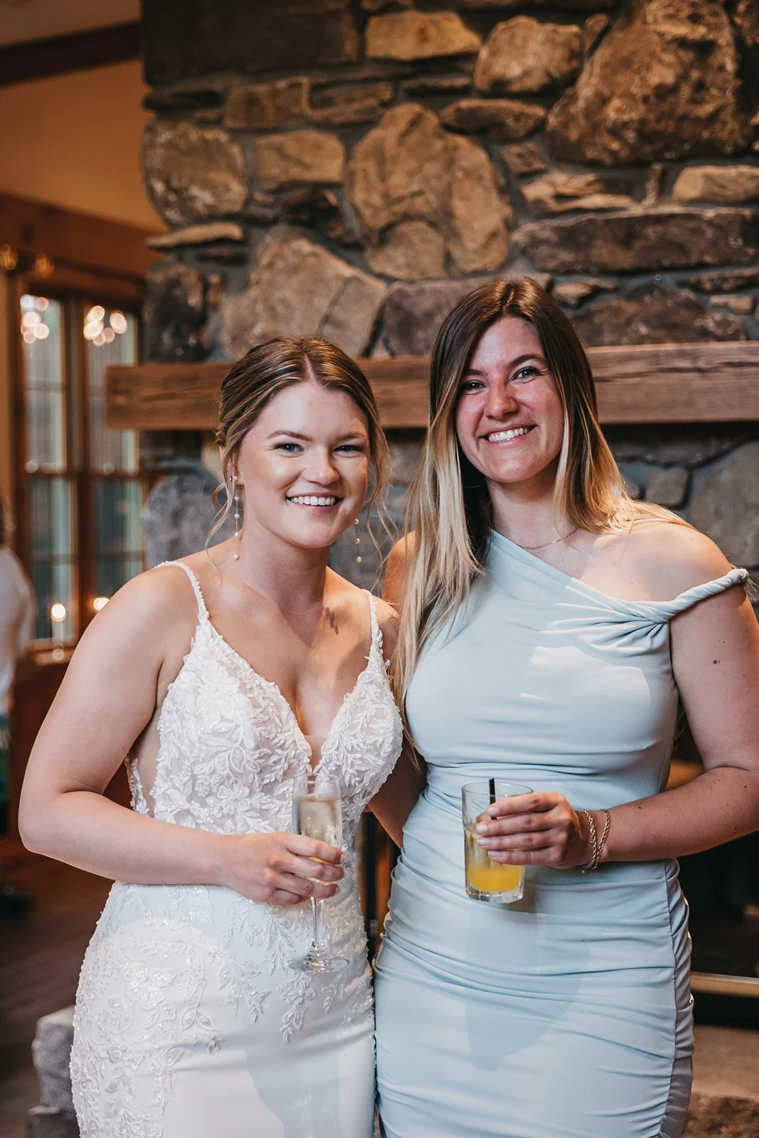 Two women smiling and holding drinks in a warmly lit room with a stone fireplace and large windows.