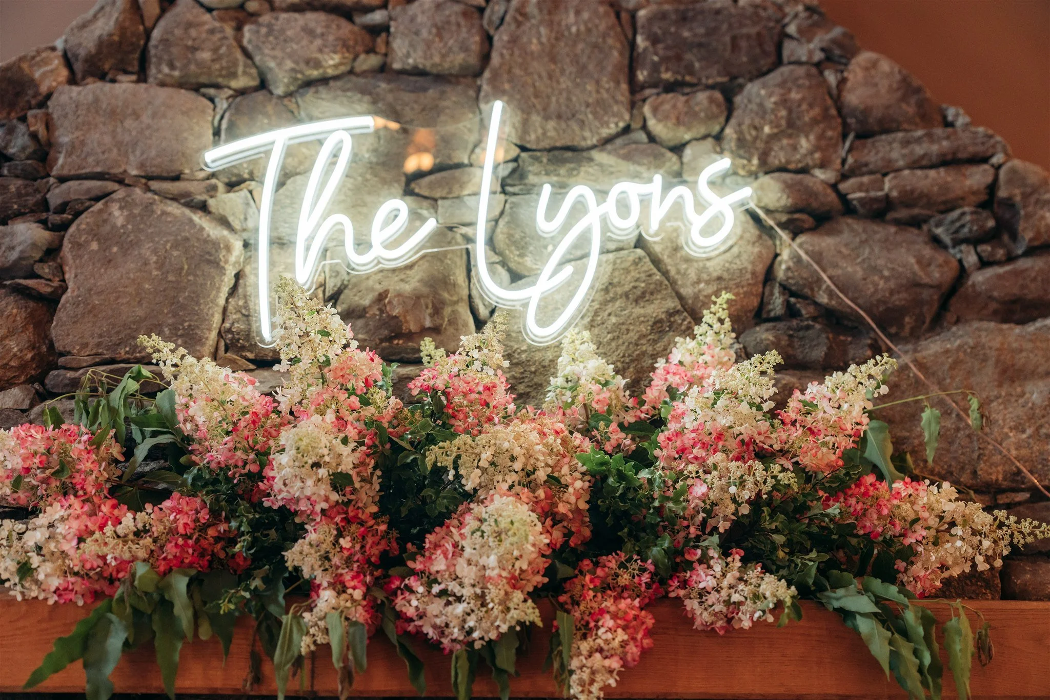 Neon sign that reads 'The Lyons' behind a large arrangement of pink and white flowers against a stone wall.