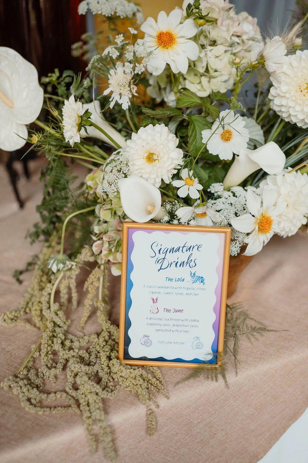 Vase with white and cream flowers. Sign on table with menu for signature drinks named The Lola and The June.