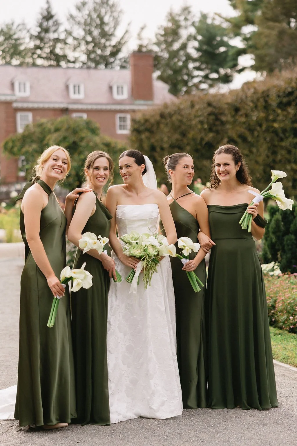 Bride in white wedding dress with four bridesmaids in dark green dresses, holding bouquets of calla lilies, outdoors with trees and a brick building in the background.