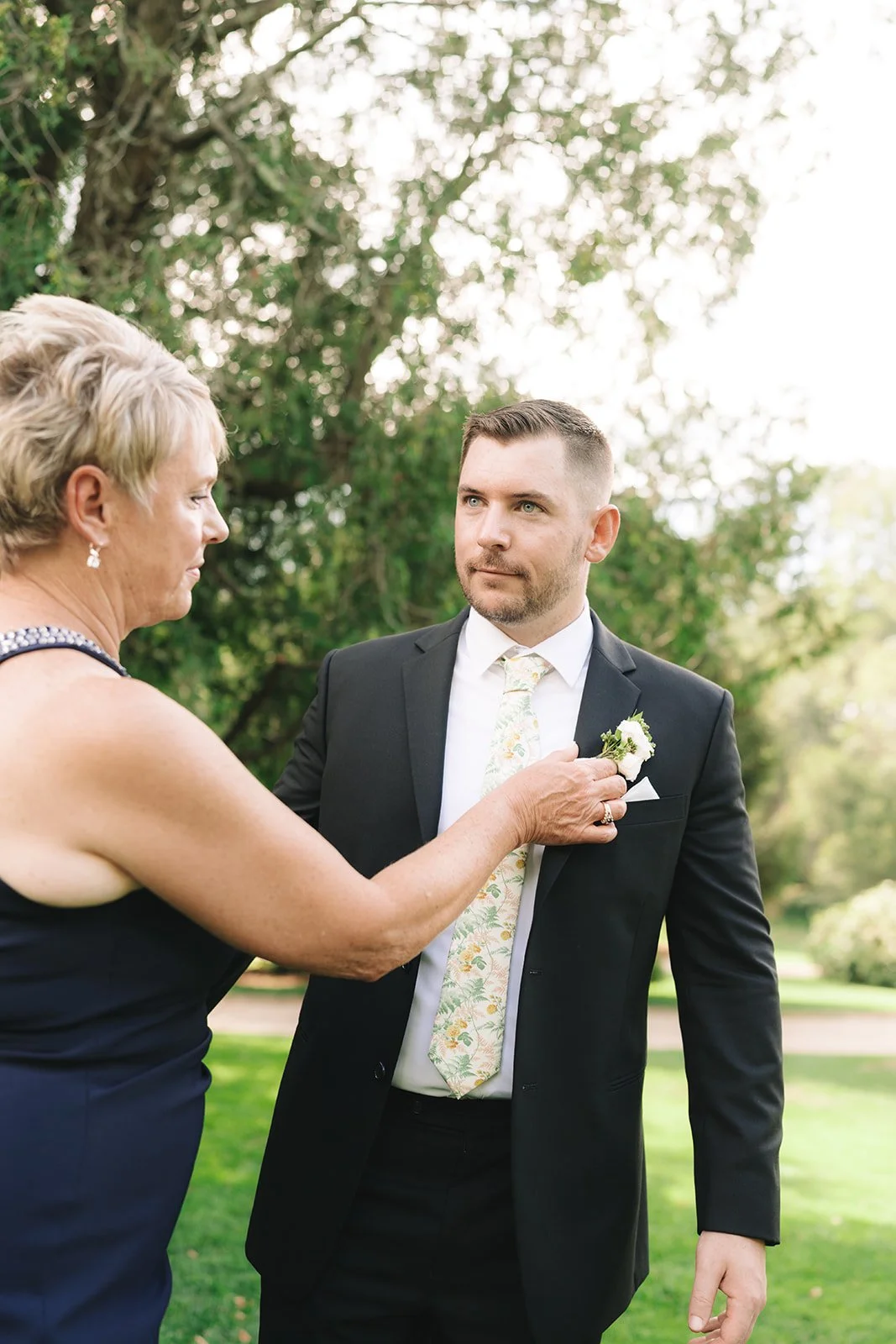 An older woman pins a boutonniere onto a young man in a black suit outside during daytime, with green trees and grass in the background.