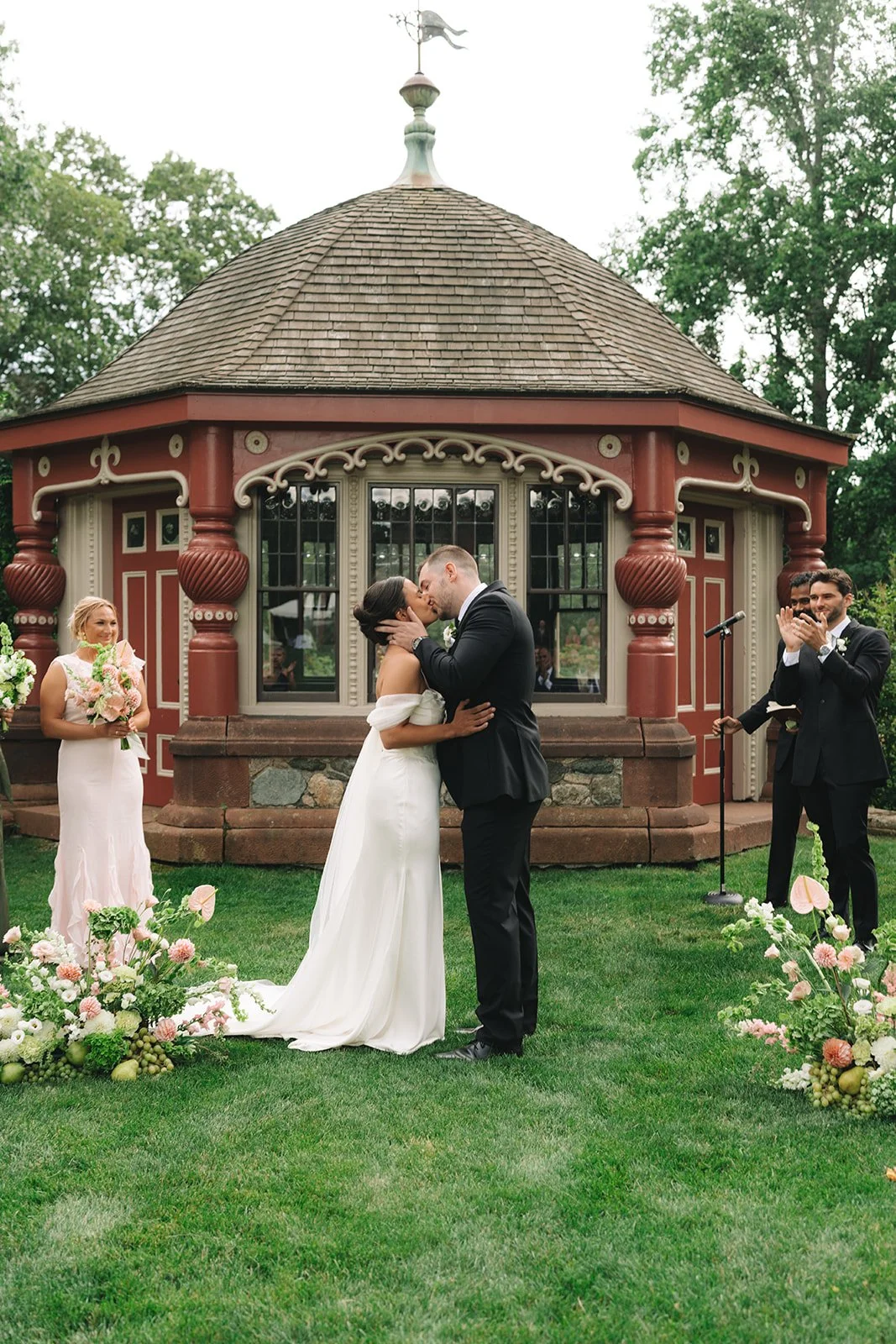 A bride and groom kiss during their wedding ceremony outdoors in front of a decorative building, with bridesmaids and groomsmen clapping and smiling nearby, surrounded by floral arrangements on green grass.