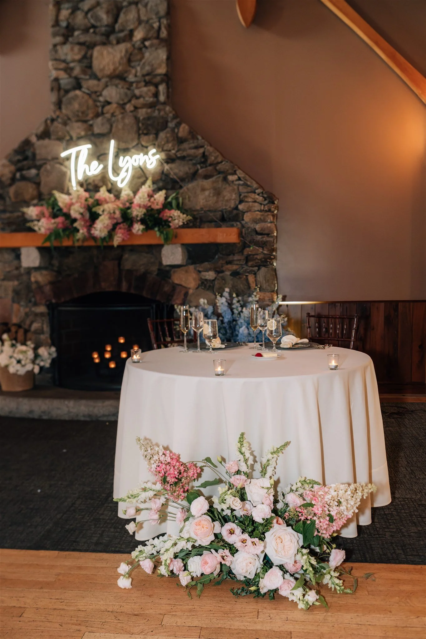 A romantic table set for a special occasion in front of a stone fireplace with pink flowers and a neon sign that reads 'The Lyons'.