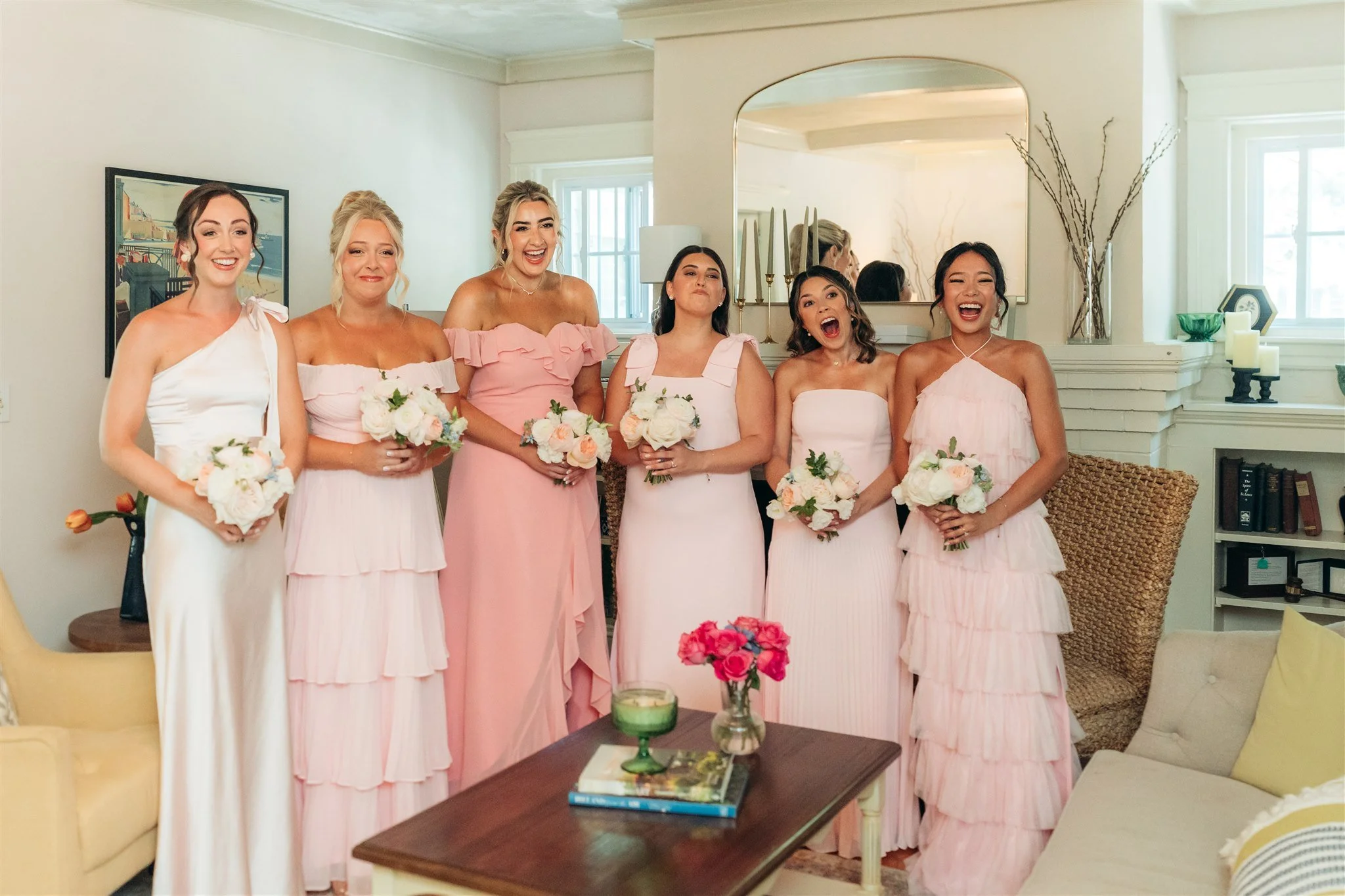 A group of seven women in pastel-colored dresses, standing inside a living room, holding bouquets of white and pink flowers, celebrating a wedding or bridal event.