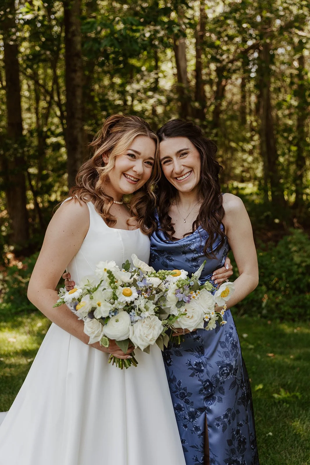 Two women standing outdoors, smiling, one in a white wedding dress holding a bouquet, the other in a blue floral dress also holding a bouquet, surrounded by trees.