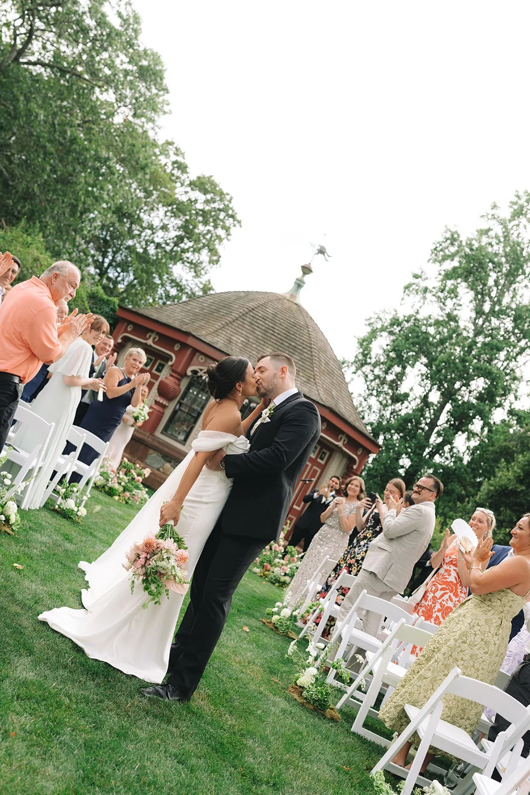 A wedding couple kisses outdoors during their ceremony with guests standing and clapping around them.