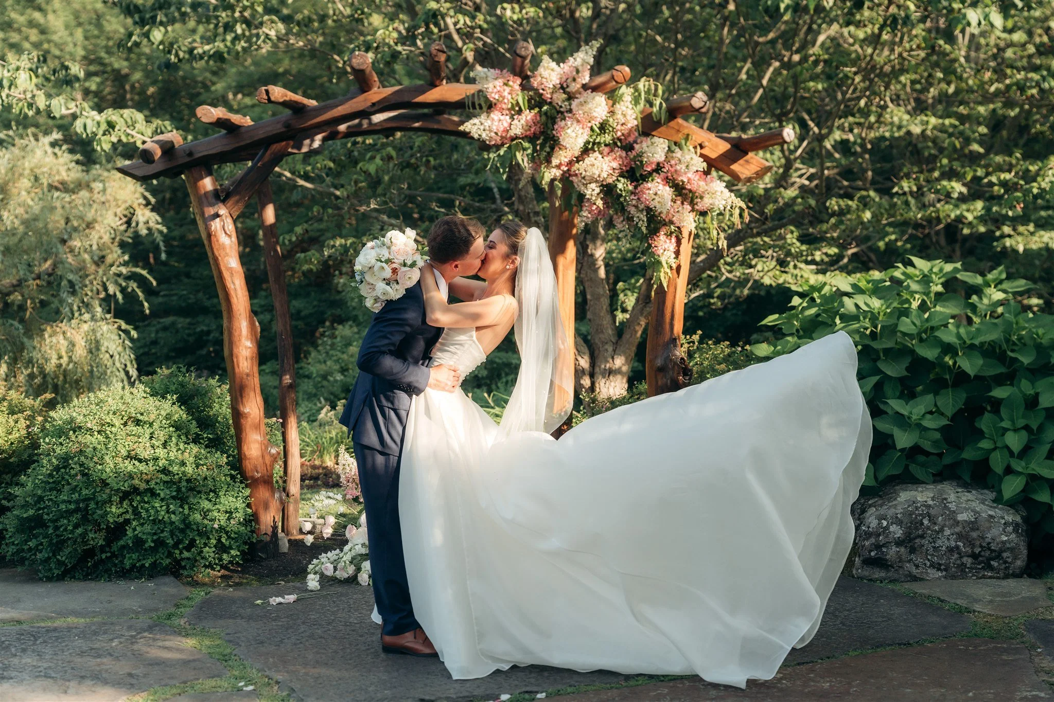 A bride and groom kissing under a wooden arch decorated with pink and white flowers during a wedding ceremony outdoors surrounded by greenery.