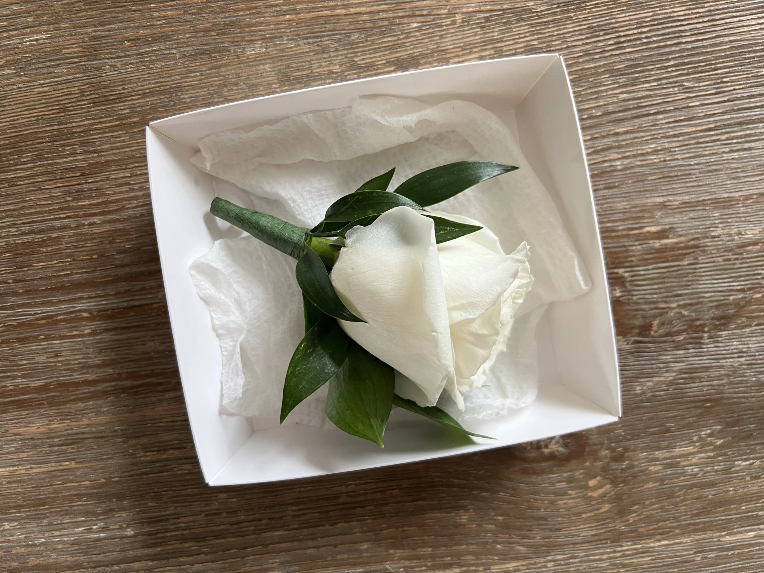 A white rose with green leaves in a white gift box on a wooden surface.